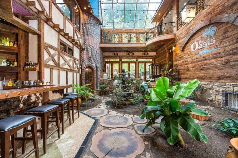 A marvelous green sunroom filled with a bar, fresh plants, and a fountain that serves as the focal visual point in the room. Aside from the natural light, magical string lights and ambient sconces illuminate the area as well. Photo Credit: Steve Haning Source: luxecir.idxbroker.com